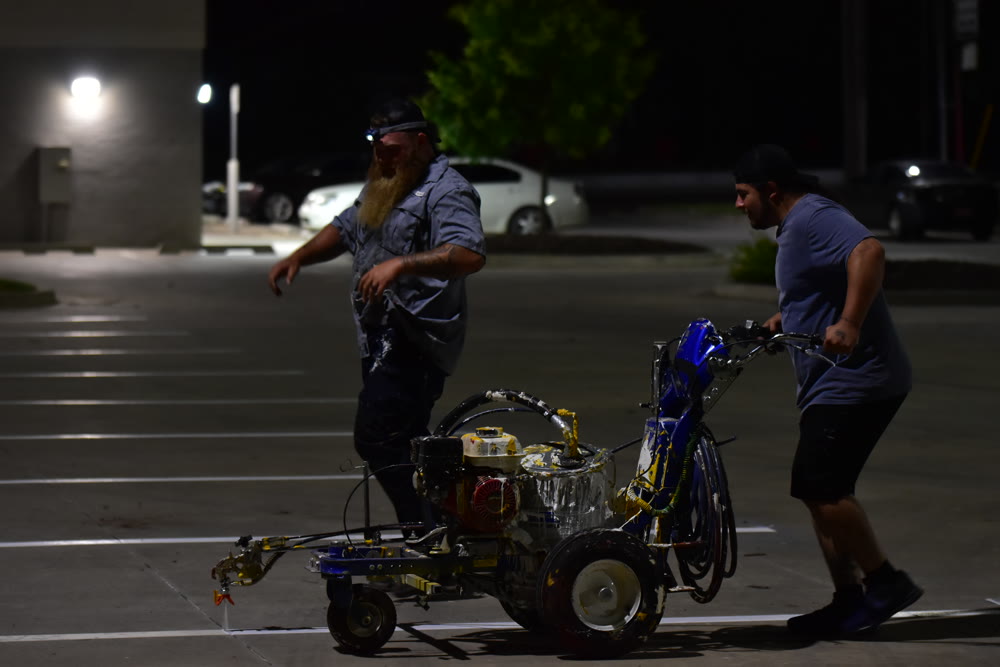 Two-man crew pushing a line striper machine across a lot at night